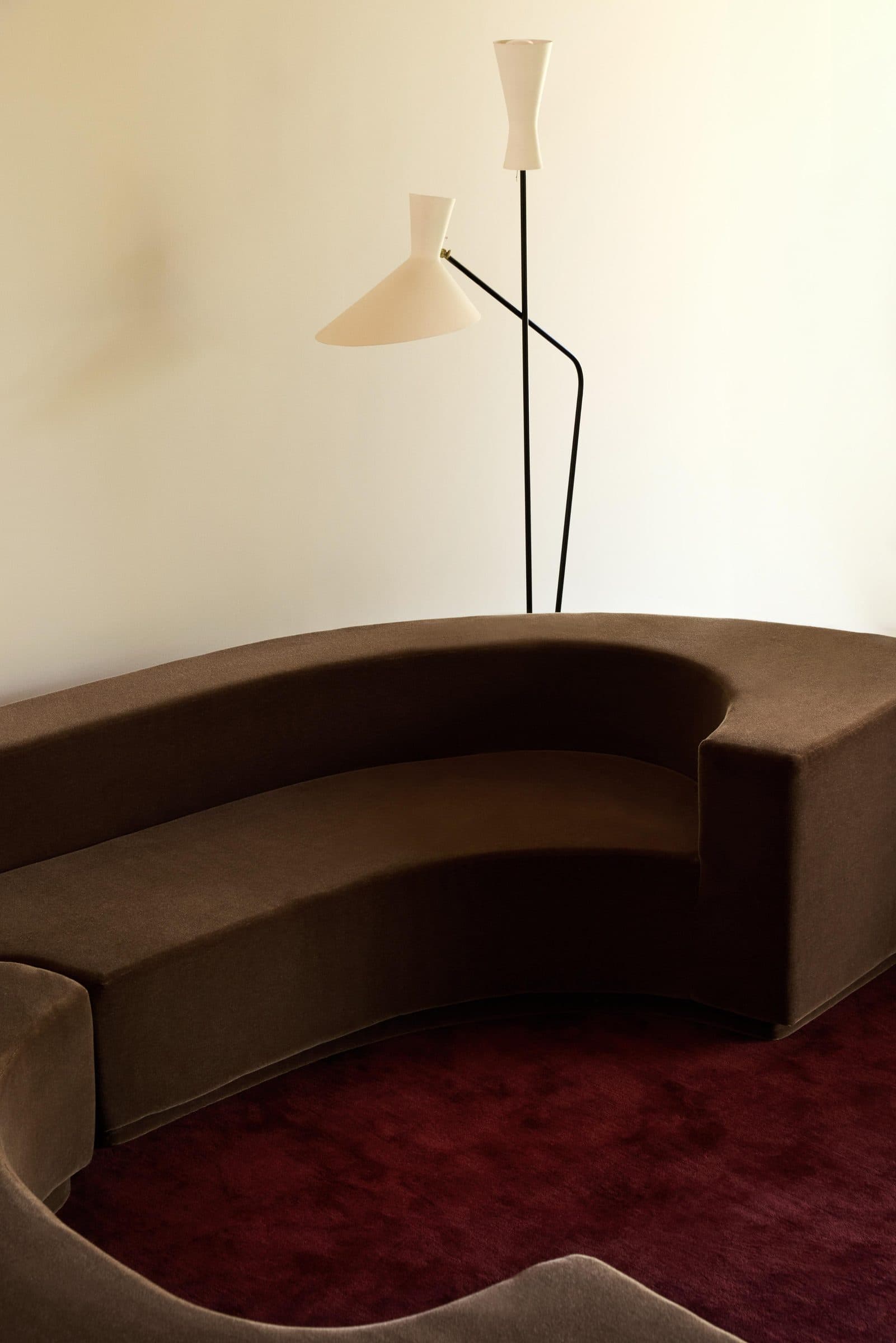 A deep brown velvet curved sofa arm against a warm cream wall, with two thin black lamp stems rising at the top.