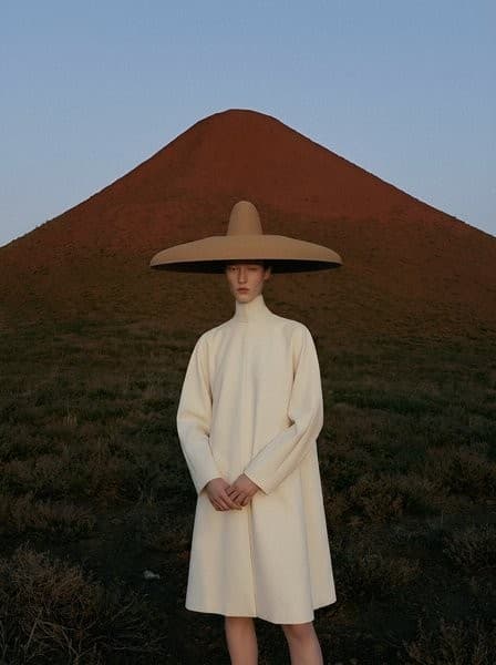 A figure in a cream coat and wide hat standing before a red hill.