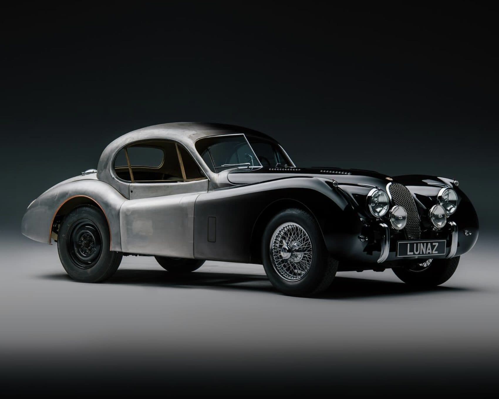 A dark grey classic sports coupe on a black studio floor.